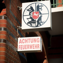 Detailaufnahme: Leuchtschilder mit Symbol „Feuerwehr“ und Aufschrift „Achtung Feuerwehr“ außen an der Fassade
