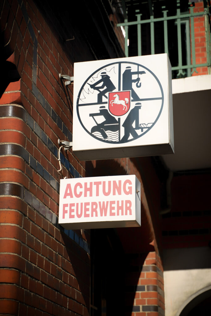 Detailaufnahme: Leuchtschilder mit Symbol „Feuerwehr“ und Aufschrift „Achtung Feuerwehr“ außen an der Fassade