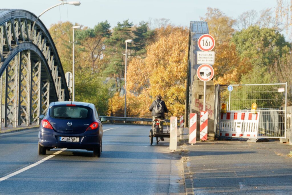 Ein Auto überholt eine Radfahrerin mit Anhänger und überfährt dabei die durchgezogene Mittellinie der Fahrbahn.