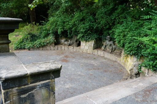 Zwei steinerne Skulpturen, die liegende Menschen zeigen, eingefasst in einer bogenförmig verlaufenden Mauer. im Vordergrund drei hinabführende Treppenstufen.